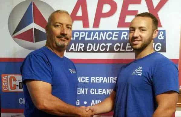 Two men in blue shirts shake hands and smile in front of a sign for Apex Appliance Repair & Air Duct Cleaning, trusted for air duct services New Jersey residents rely on.