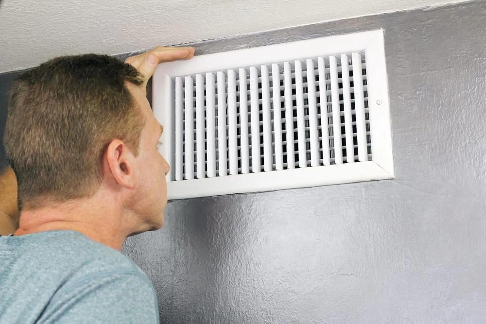 A man in a blue shirt inspects a white air vent on a gray wall, looking closely at the grille&mdash;a scene reflecting the thorough attention given during professional air duct services in New Jersey.