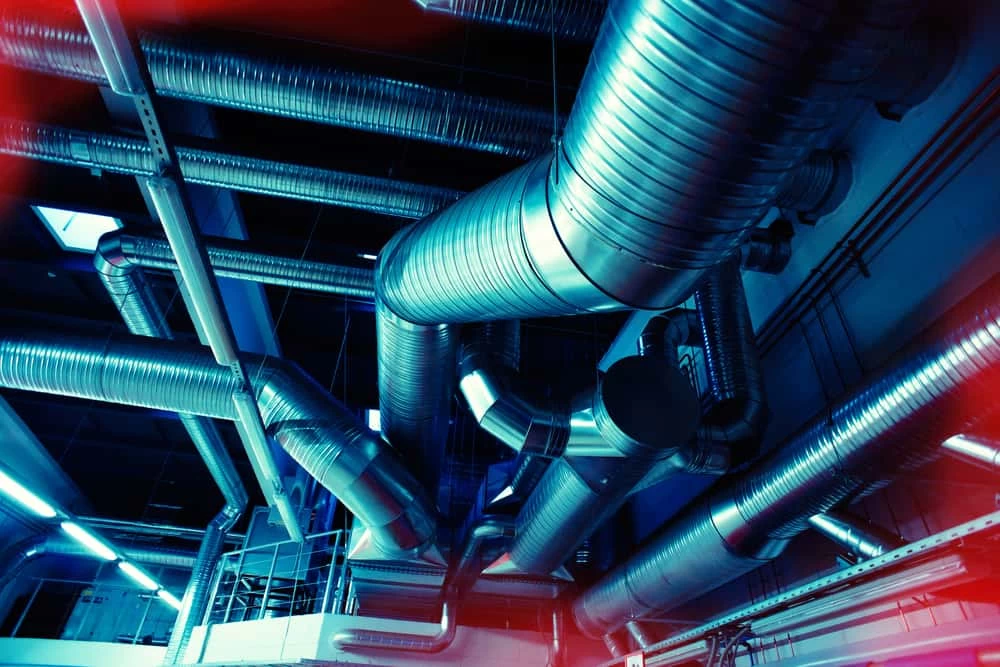 View of an industrial ceiling filled with large, shiny metal air ducts and ventilation pipes, illuminated by blue-toned lighting with red light flares&mdash;an impressive showcase for professional air duct services New Jersey offers.
