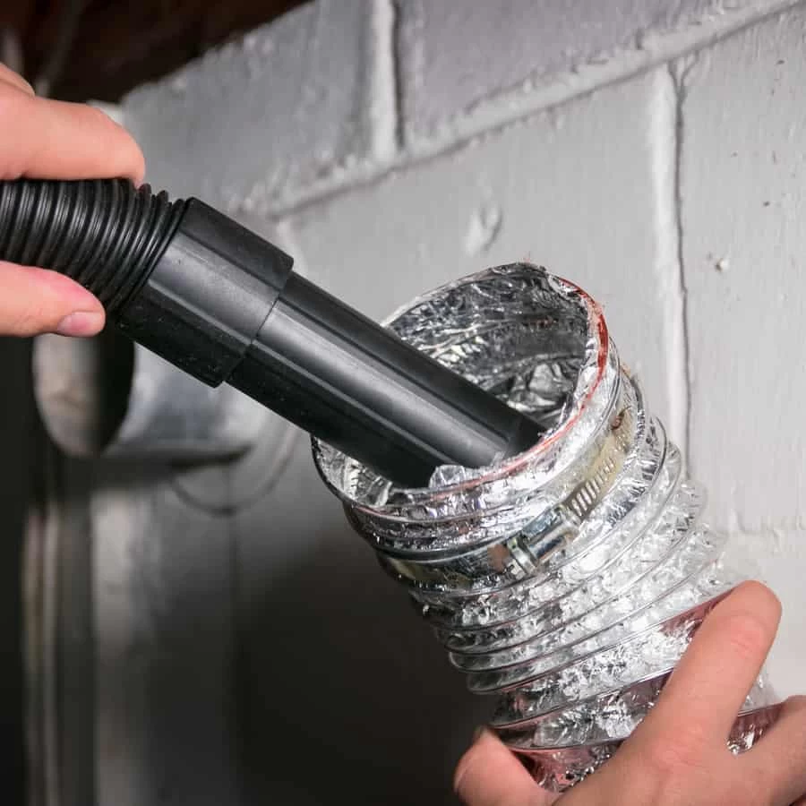 A person uses a vacuum hose to clean the inside of a flexible, metal dryer vent duct attached to a wall, demonstrating professional dryer vent cleaning New Jersey homeowners can trust.