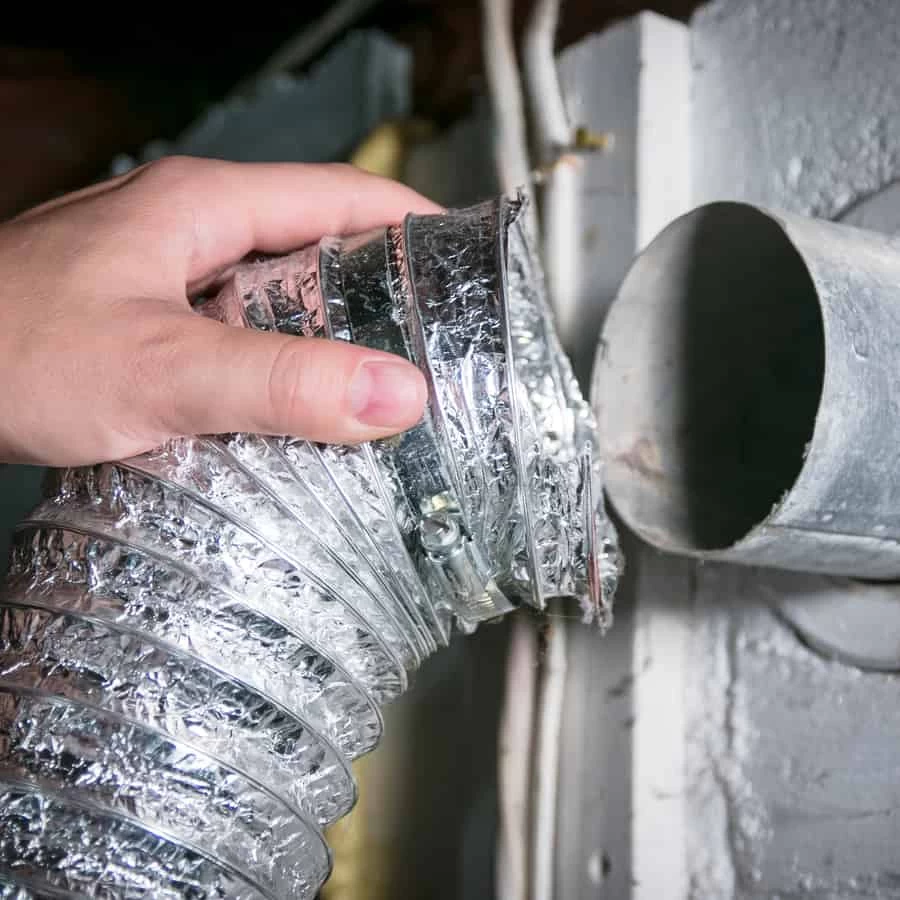 A hand connects a flexible, silver foil dryer vent hose to a round metal exhaust duct on a wall, highlighting the importance of regular dryer vent cleaning New Jersey homeowners need for safety and efficiency.