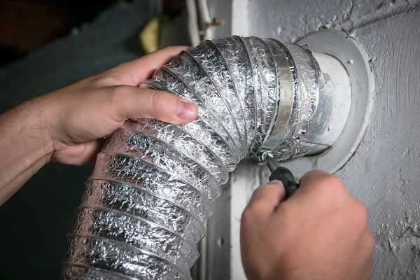 Close-up of hands attaching a flexible silver dryer vent hose to a wall vent using a screwdriver, securing the hose clamp in place&mdash;part of professional dryer vent cleaning New Jersey homeowners trust.