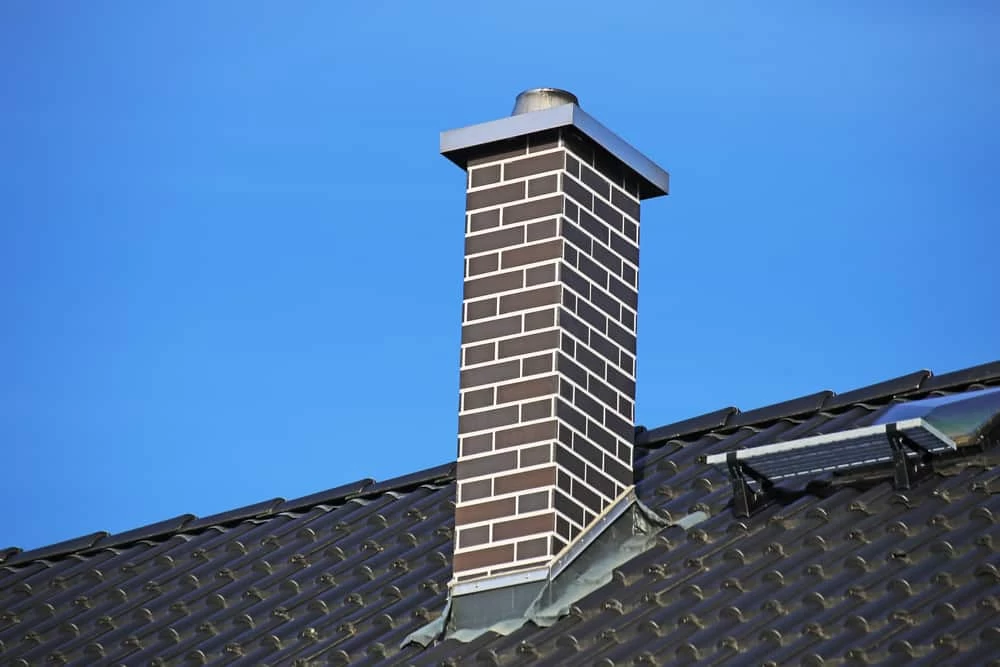 A brick chimney with a metal cap rises above a dark tiled roof, set against a clear blue sky. Part of a skylight or solar panel is visible on the right side&mdash;perfect for homes needing dryer vent cleaning New Jersey can trust.