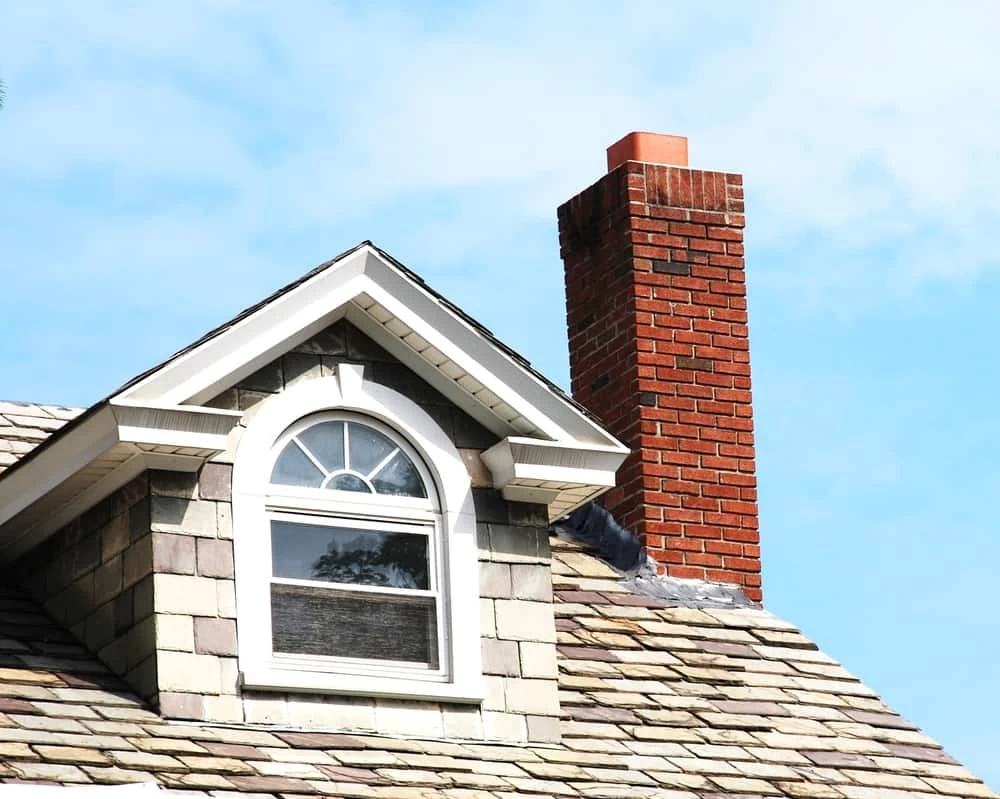 A brick chimney rises above a sloped shingle roof with a small arched window dormer, set against a blue sky with light clouds&mdash;perfect inspiration for homes seeking expert dryer vent cleaning New Jersey trusts.