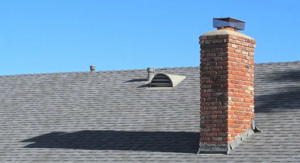 A red brick chimney rises from a gray shingled roof under a clear blue sky, with a metal vent and small exhaust pipe&mdash;ideal spots for dryer vent cleaning New Jersey professionals to inspect&mdash;also visible on the roof.