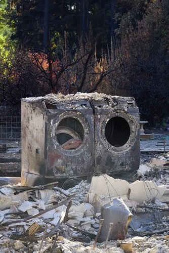 Two heavily damaged, soot-covered washing machines stand amid debris and rubble, with scorched trees and burnt surroundings in the background, highlighting the importance of dryer vent cleaning New Jersey after a destructive fire.
