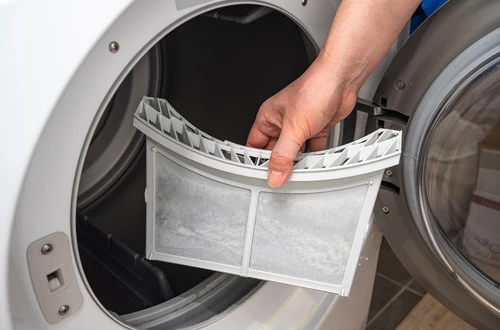 A person&rsquo;s hand removes a lint filter covered in dust from the open door of a front-loading dryer, emphasizing the importance of regular dryer vent cleaning New Jersey to maintain safety and efficiency.
