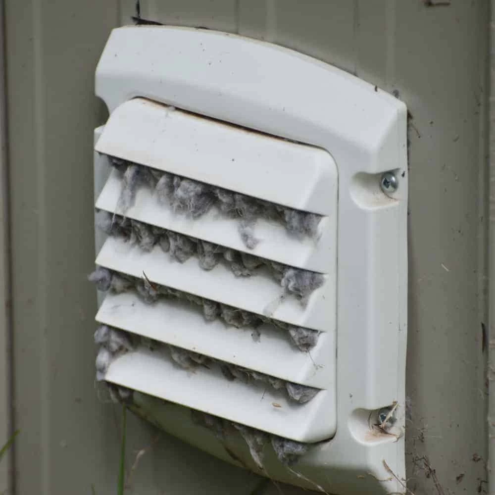 A white exterior vent cover, clogged with gray lint and debris, is attached to the side of a building&mdash;a clear sign that dryer vent cleaning New Jersey might be needed.