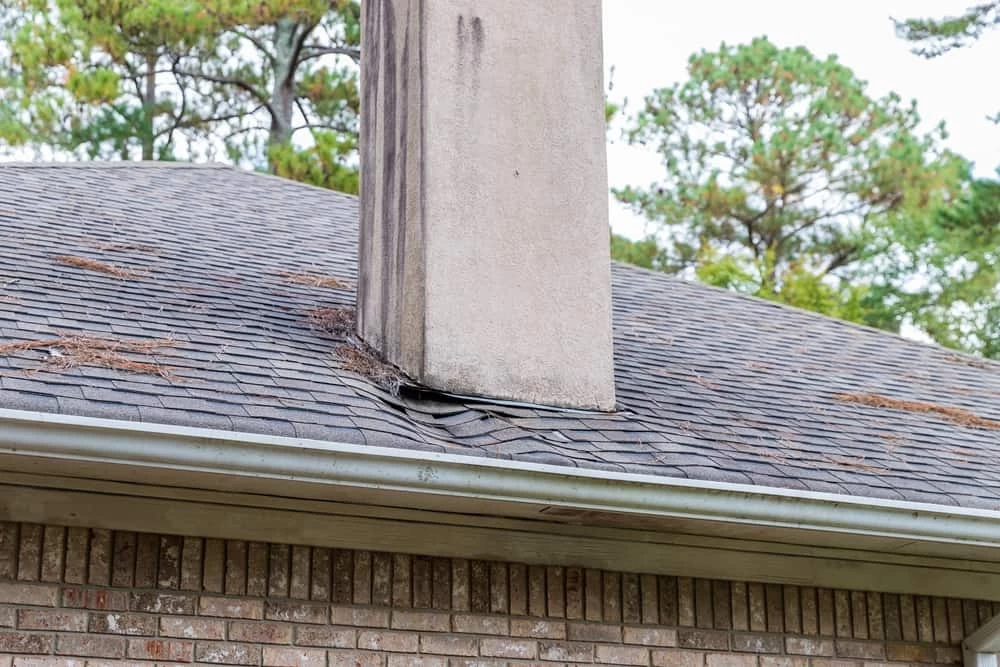 A house roof with missing shingles and heavy wear around a chimney, along with debris and pine needles scattered on the shingles. Trees are visible in the background&mdash;an ideal reminder to consider air duct services New Jersey for overall home care.