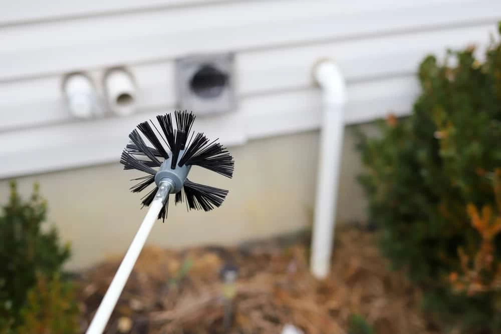 A close-up of a round dryer vent cleaning brush held outside, with a house wall, dryer vent, and some bushes visible in the background&mdash;perfect for showcasing expert dryer vent cleaning New Jersey services.