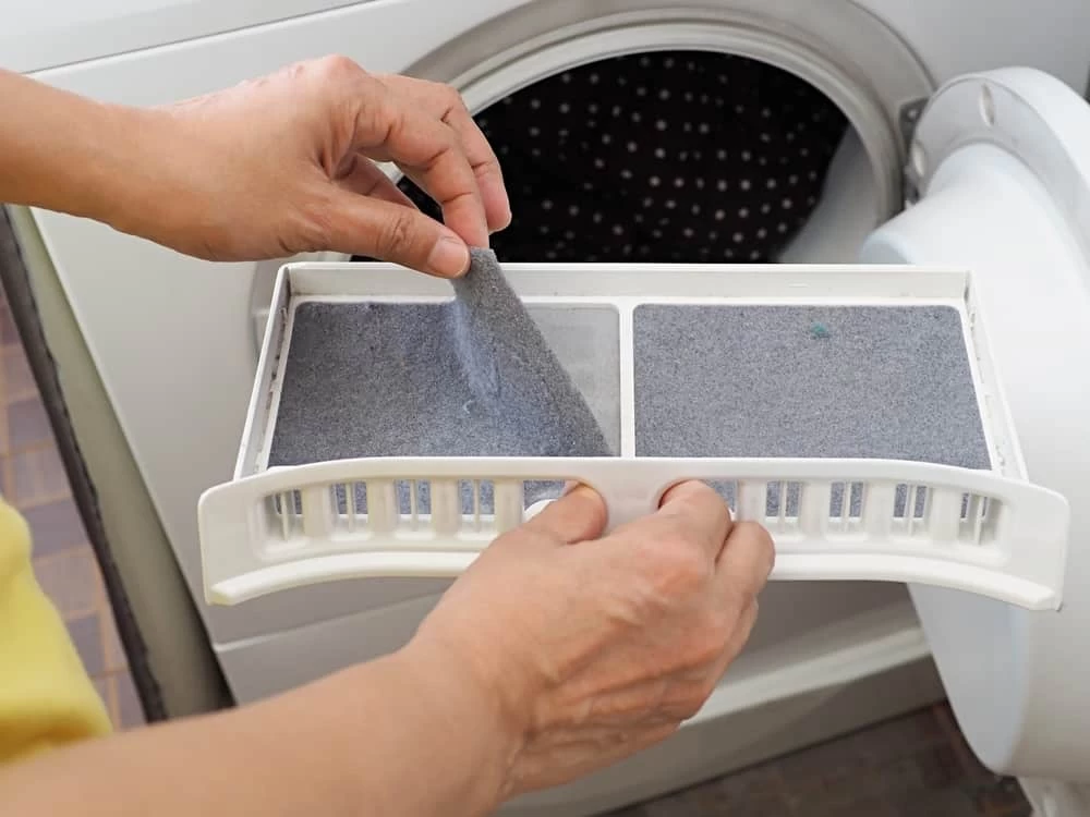 A person is removing lint from a filter tray of a front-loading dryer, with the dryer door open and clothes visible inside&mdash;essential for safe operation and recommended by dryer vent cleaning New Jersey professionals.