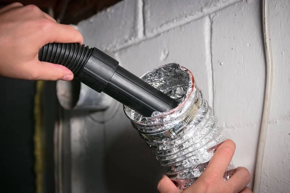 A person holds a vacuum hose inside a flexible dryer vent duct, cleaning it near a white painted brick wall&mdash;demonstrating professional dryer vent cleaning New Jersey services.