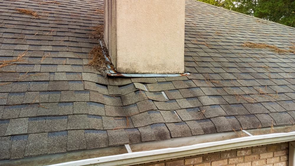 A shingled roof with a chimney shows damaged, warped shingles and poor flashing. Pine needles are scattered on the roof, and a white rain gutter runs along the edge above a brick wall&mdash;perfect for noting dryer vent cleaning New Jersey needs.