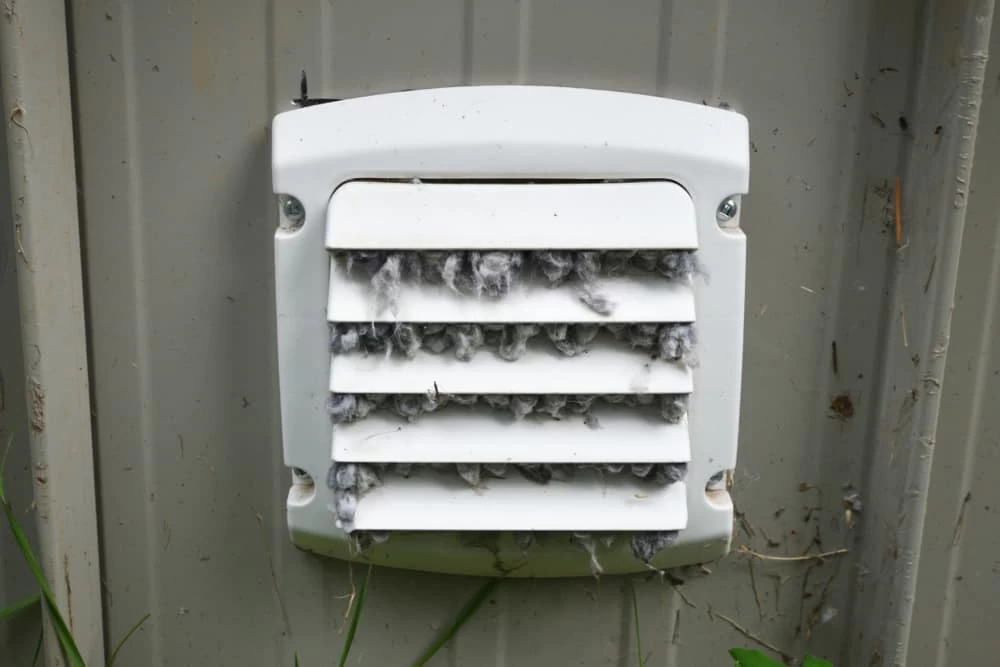 A white exterior vent cover, partially clogged with lint and debris, is attached to a light gray wall with green grass below&mdash;a clear sign that dryer vent cleaning New Jersey homeowners need could improve airflow and safety.