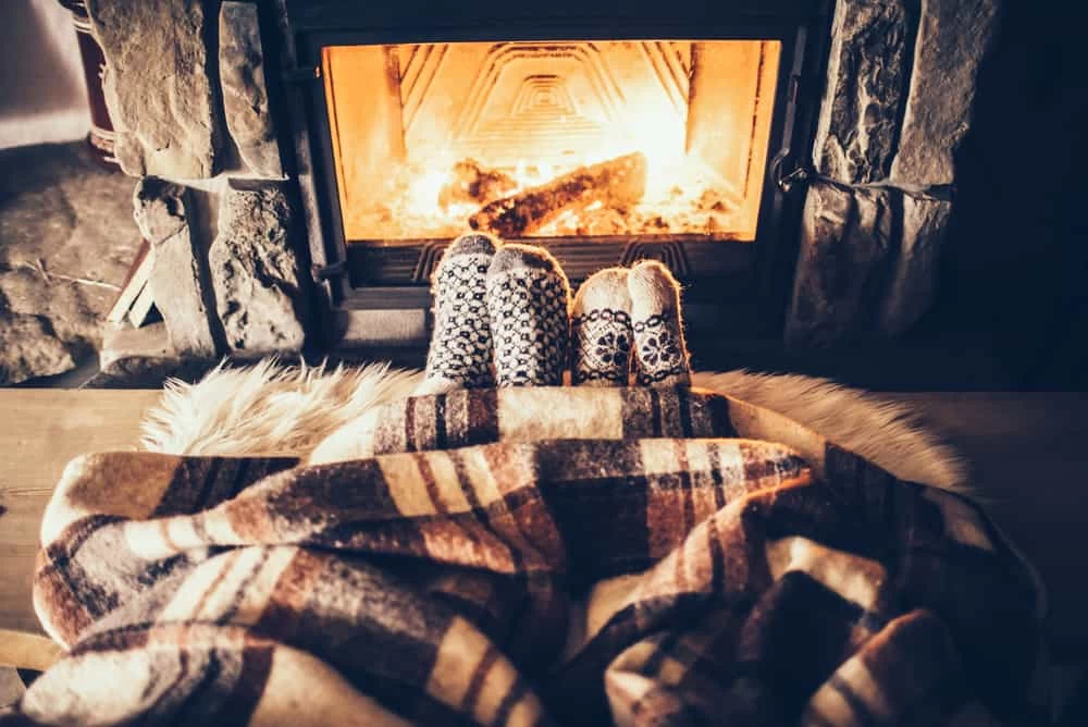 Two people wearing patterned socks relax by a burning fireplace, covered by a plaid blanket and fur rug, creating a cozy vibe&mdash;perfect after scheduling dryer vent cleaning New Jersey to keep your home safe and warm all winter.