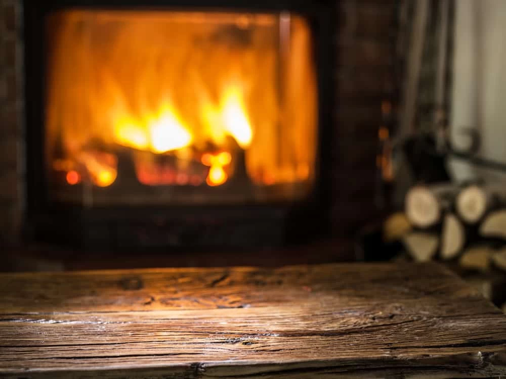 A close-up of a rustic wooden table in front of a glowing fireplace with burning logs, creating a warm and cozy atmosphere. Stacked firewood is visible on the right side, reminding homeowners about the importance of dryer vent cleaning New Jersey.