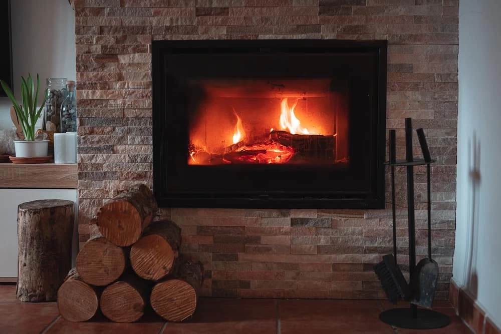 A cozy fireplace with burning logs is set into a stone wall. Firewood is stacked neatly beside it, along with fireplace tools and a small potted plant on a nearby shelf&mdash;perfect for relaxing after air duct services New Jersey.