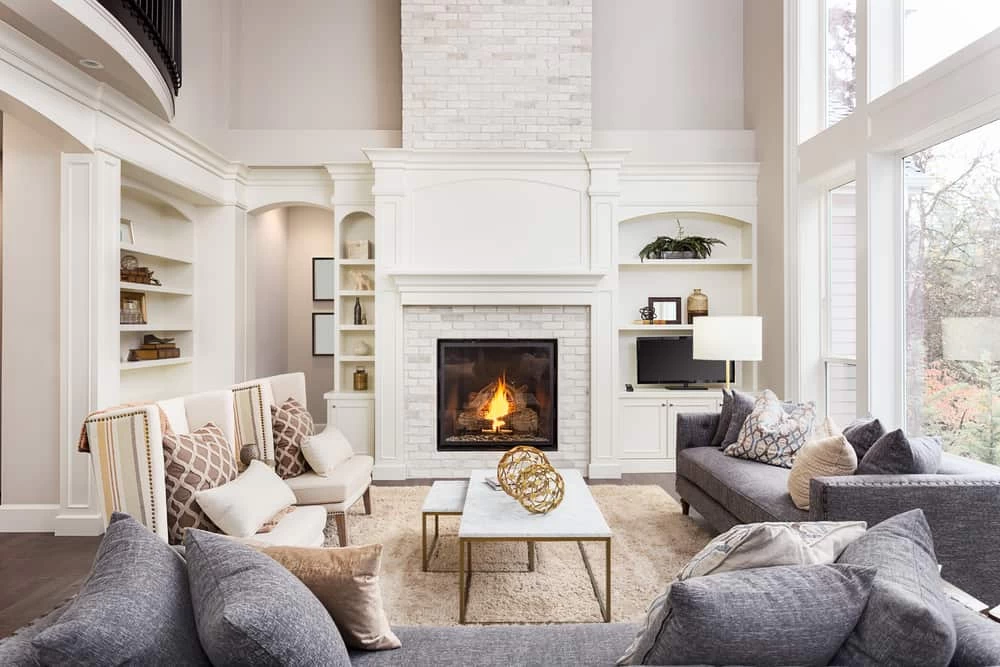 Bright, elegant living room with a lit fireplace, gray sofas, cushioned armchairs, a marble coffee table, built-in shelves, large windows, and neutral decor&mdash;enhanced by fresh air from professional air duct services New Jersey.