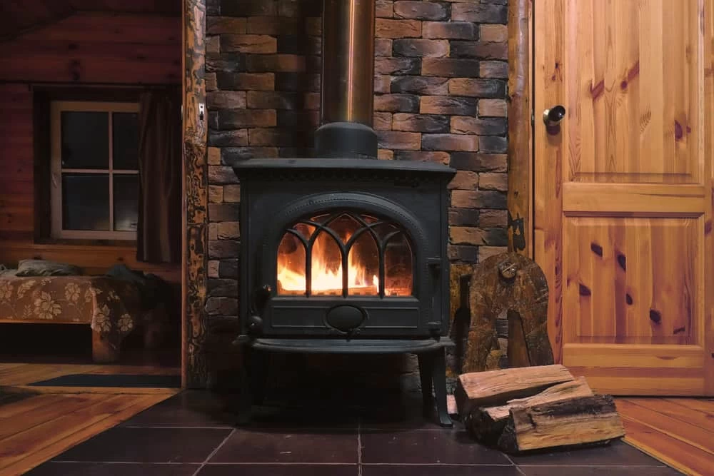 A black wood-burning stove with a fire inside sits on a tiled floor in front of a brick wall, with logs stacked beside it in a cozy wooden cabin interior, highlighting the importance of air duct services New Jersey for maintaining indoor air quality.