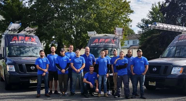 A group of thirteen people in blue shirts stand in front of two black vans labeled "Apex Air Duct Cleaning," parked on a residential street, ready to provide top-notch air duct services New Jersey homeowners can trust.