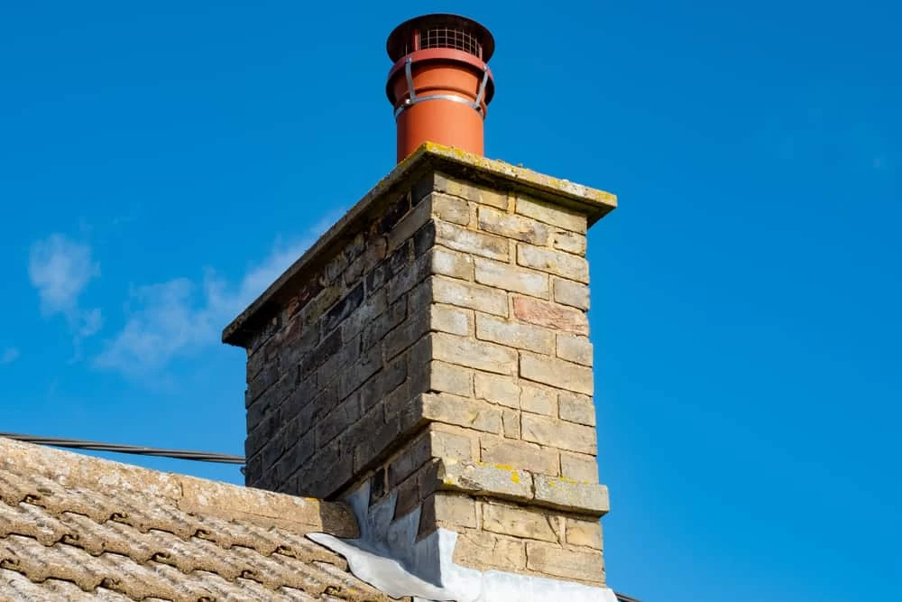 A close-up of a brick chimney with a red metal cap, set against a clear blue sky, on the roof of a house with gray shingles&mdash;ideal for showcasing expert dryer vent cleaning New Jersey homeowners can trust.