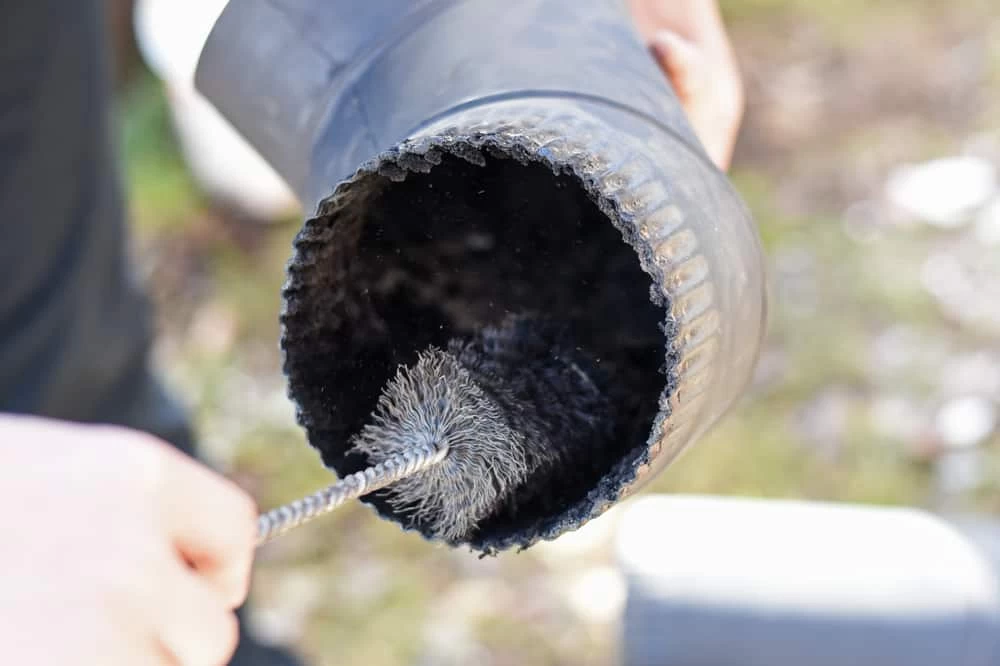 A person cleans the inside of a large black pipe with a round wire brush tool, focusing on removing soot or debris&mdash;similar to professional dryer vent cleaning New Jersey specialists provide.