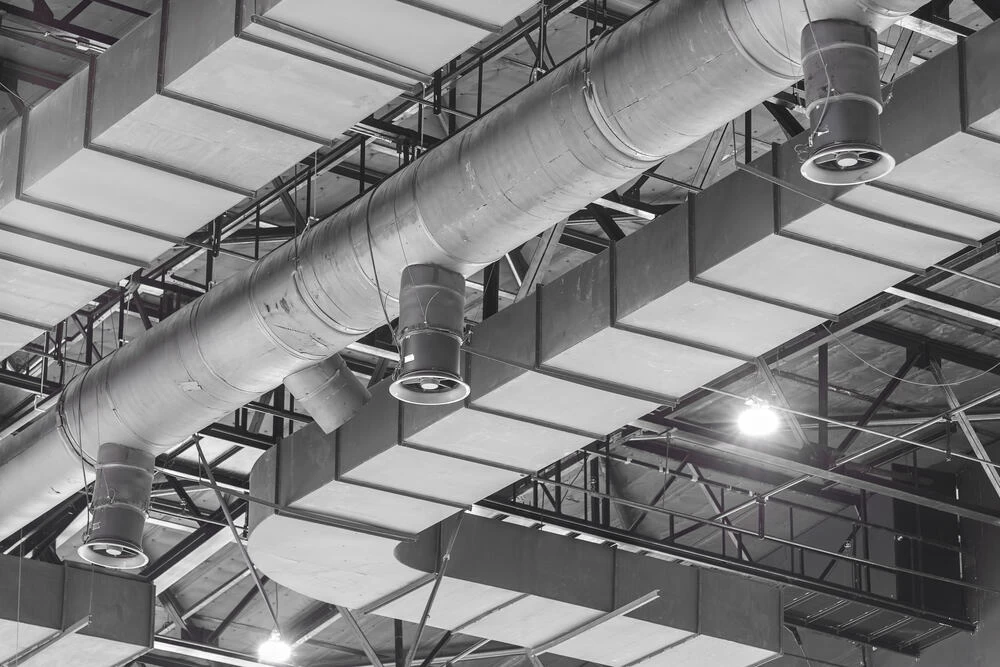 Black and white photo of an industrial ceiling showing exposed metal air ducts&mdash;perfect for showcasing professional air duct services New Jersey&mdash;along with suspended rectangular panels, structural beams, and ceiling lights.