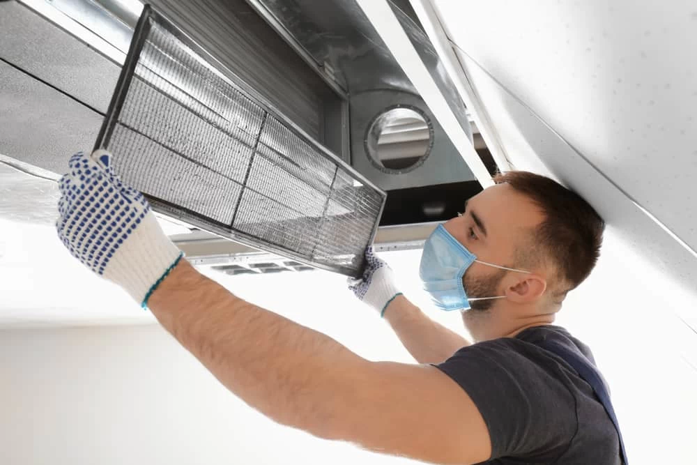 A man wearing gloves and a face mask cleans or replaces an air filter in a ceiling-mounted HVAC unit, showcasing professional air duct services New Jersey residents trust for cleaner indoor air.