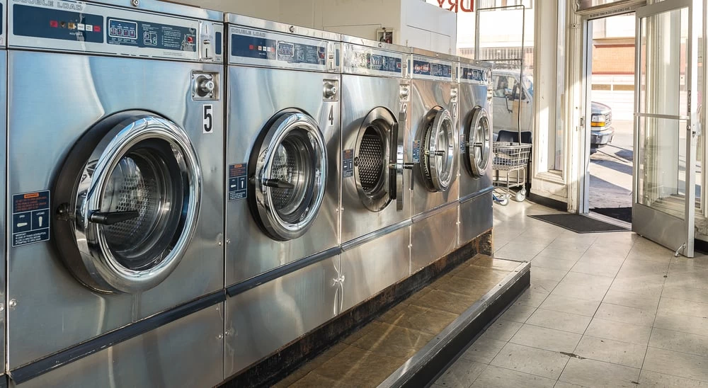 A row of industrial front-loading washing machines inside a bright laundromat. Sunlight streams through large windows, and an open glass door leads outside. Air duct services New Jersey help keep the fresh air circulating in such clean spaces. A laundry cart is visible in the background.