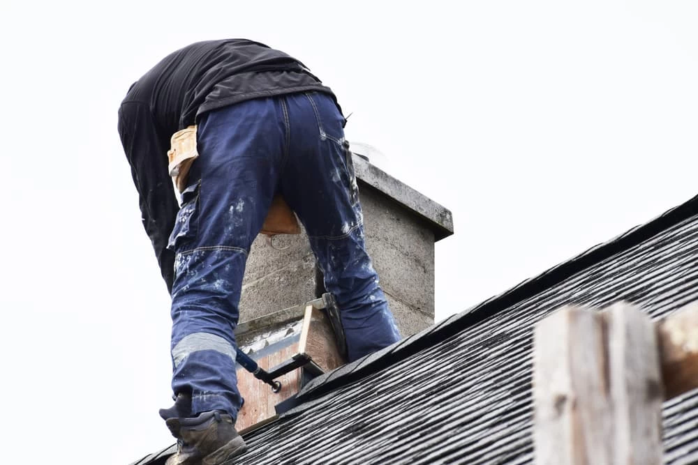A worker in paint-splattered pants stands on a sloped roof near a chimney, possibly performing dryer vent cleaning New Jersey, using tools with their back facing the camera against a white sky.