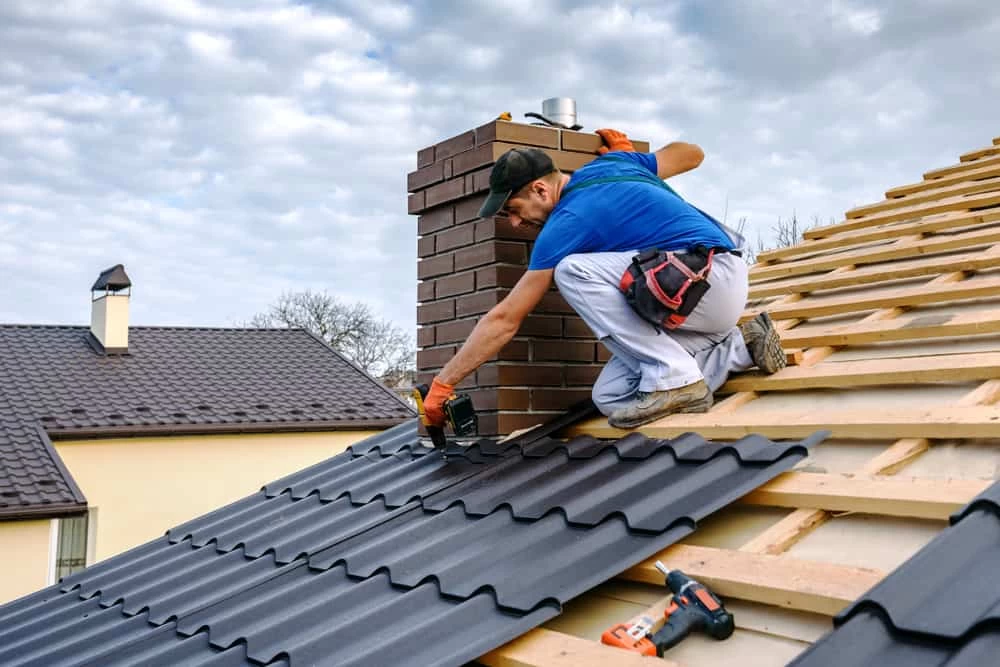 A construction worker wearing gloves and a tool belt installs dark metal roofing sheets on a wooden roof frame near a brick chimney under a cloudy sky, highlighting the attention to detail found in dryer vent cleaning New Jersey experts provide. A power drill is nearby on the roof.