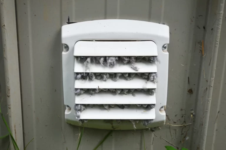 A white exterior vent cover with horizontal slats is clogged with gray lint and debris, attached to a beige wall with some grass below&mdash;reminding homeowners of the importance of regular dryer vent cleaning New Jersey.