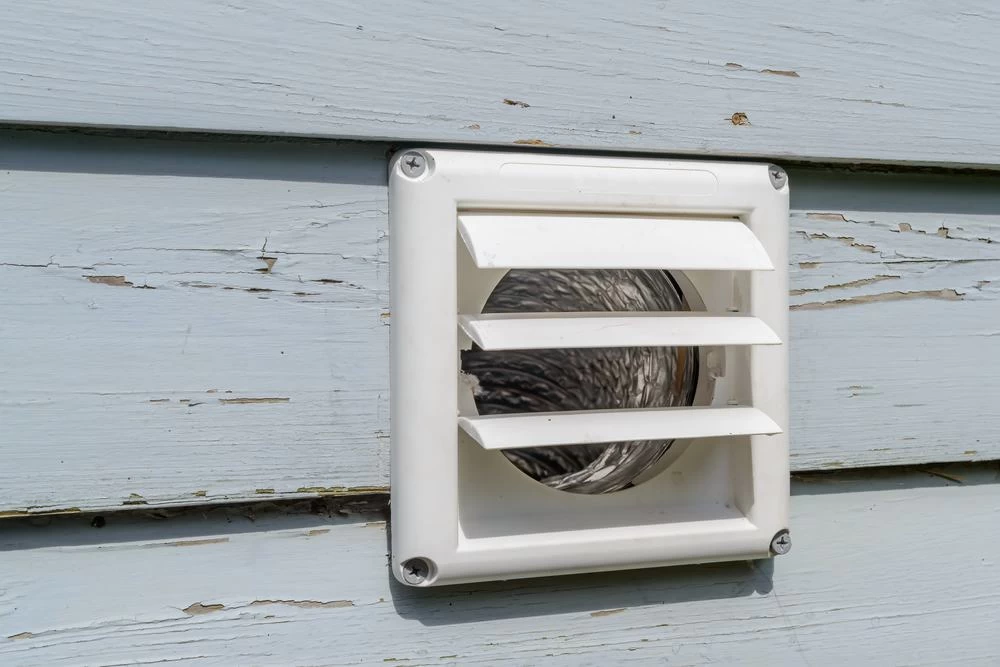 A close-up of a white vent cover with horizontal slats attached to a light blue wooden exterior wall, showing some peeling paint and weathering&mdash;a reminder of the importance of regular dryer vent cleaning New Jersey homeowners need.