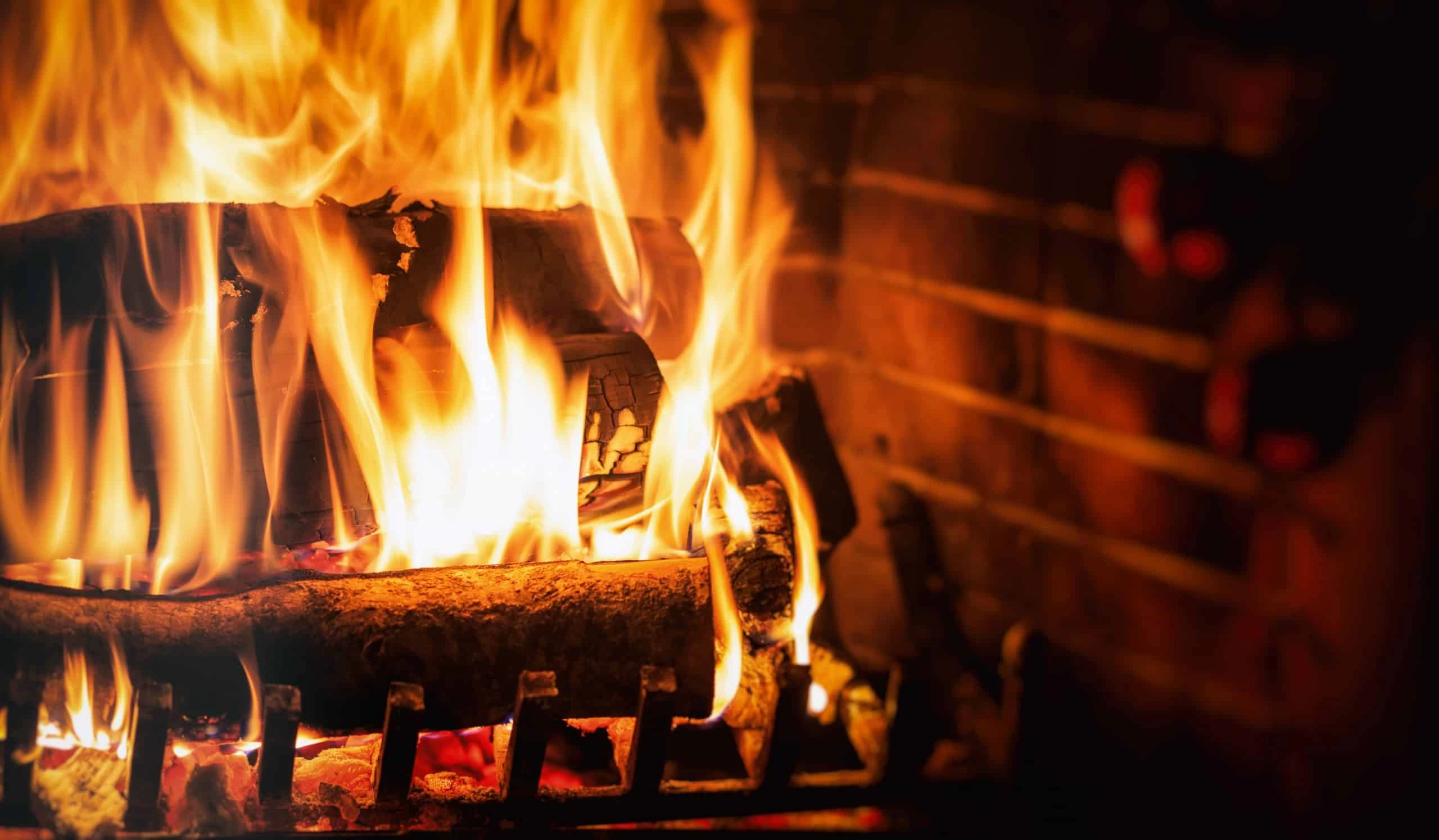 Close-up of a burning fireplace with bright orange and yellow flames consuming wooden logs, set against a brick backdrop and metal grate&mdash;reminding you of the importance of dryer vent cleaning New Jersey for home safety and warmth.