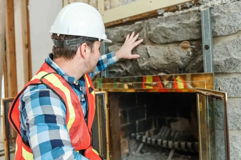 A construction worker in a hard hat and safety vest inspects or installs stones above a fireplace, with one hand touching the stone surface and the fireplace doors open&mdash;similar to professionals providing air duct services New Jersey homeowners trust.