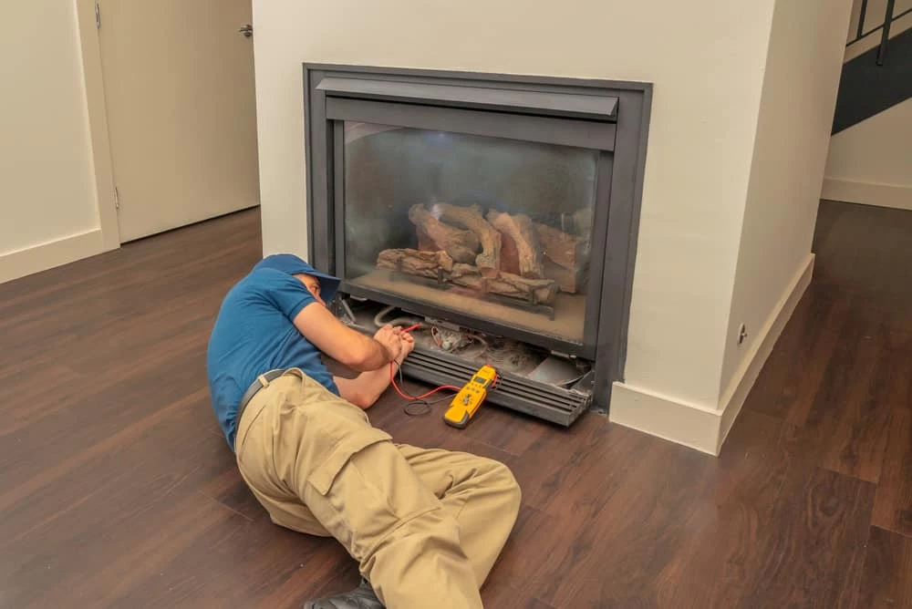A technician in a blue shirt and khaki pants is lying on the floor in front of a gas fireplace, using tools for maintenance or repairs&mdash;typical of top dryer vent cleaning New Jersey professionals. A yellow multimeter rests nearby on the floor.