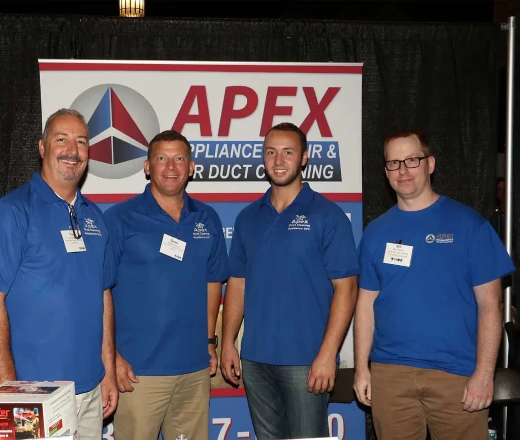 Four men in blue shirts and name tags stand in front of an "APEX Appliance Repair & Air Duct Cleaning" booth, smiling at the camera&mdash;experts in air duct services New Jersey and dryer vent cleaning New Jersey.