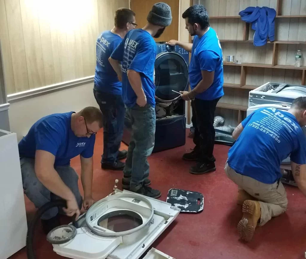 Four men in blue shirts work together to repair washing machines in a room with wooden walls and shelves. Parts and tools are spread out on the red floor as they focus on tasks, showcasing their expertise in dryer vent cleaning New Jersey.