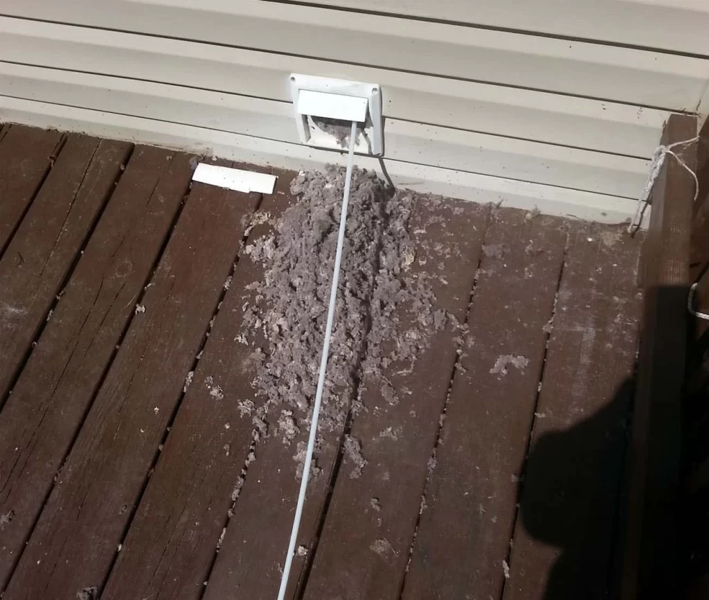 A pile of gray lint scattered on a brown wooden deck beneath an exterior dryer vent&mdash;reminding homeowners of the importance of regular dryer vent cleaning New Jersey, with a white vent cover and shadow visible on the right.