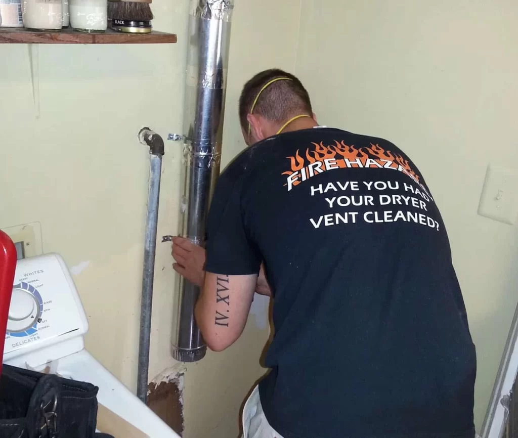 A man in a black &ldquo;FIRE HAZARD: HAVE YOU HAD YOUR DRYER VENT CLEANED?&rdquo; shirt is cleaning a metal dryer vent in a laundry room, showcasing professional dryer vent cleaning New Jersey services next to a washing machine.