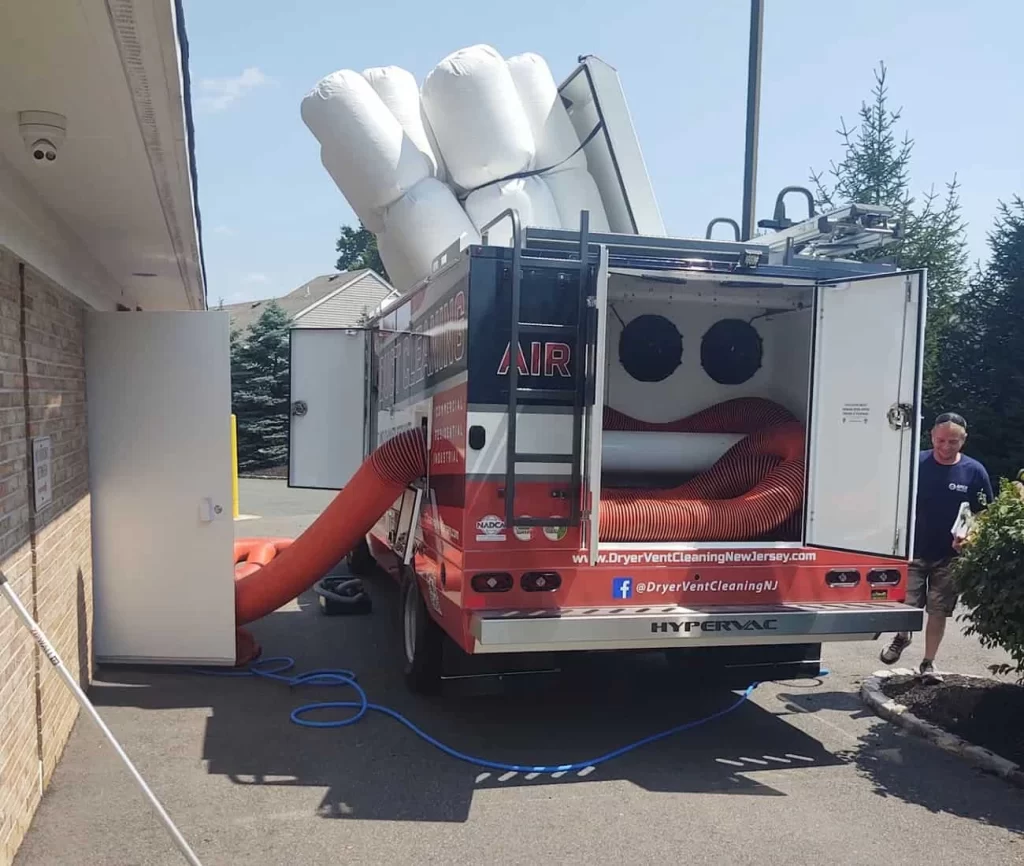 A large cleaning truck with open rear doors uses big orange hoses to connect to a building for air duct services New Jersey. White inflatable bags are attached to the top. A man stands nearby on the right side.