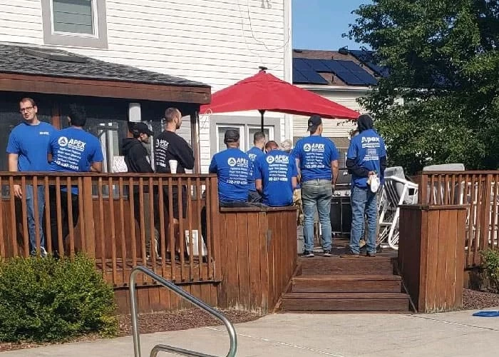A group of people wearing matching "APEX" shirts, representing air duct services New Jersey, stand and sit on a wooden deck outside a house with white siding. A red umbrella and patio furniture are visible in the background.