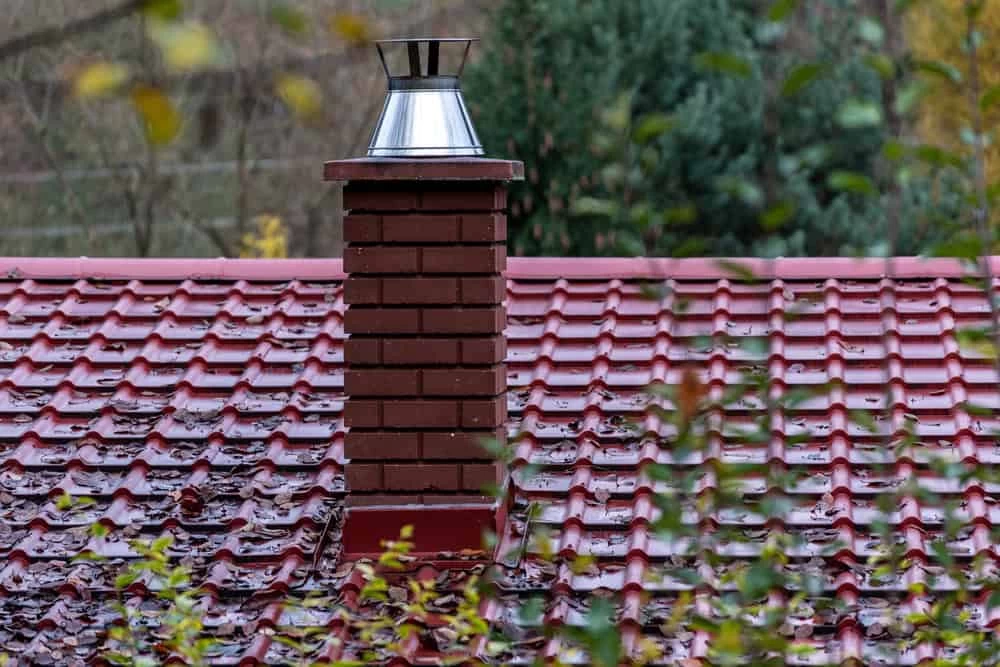 A brick chimney with a metal cap rises from a red, tiled roof scattered with fallen leaves, surrounded by leafy green trees&mdash;an ideal setting to consider air duct services New Jersey for a cleaner and safer home environment.