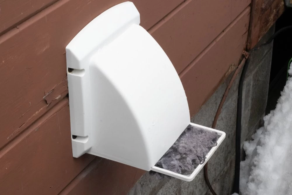 A white dryer vent cover is mounted on the side of a brown building, with a tray beneath it catching lint&mdash;a reminder of the importance of regular dryer vent cleaning New Jersey. Snow is visible on the ground nearby.