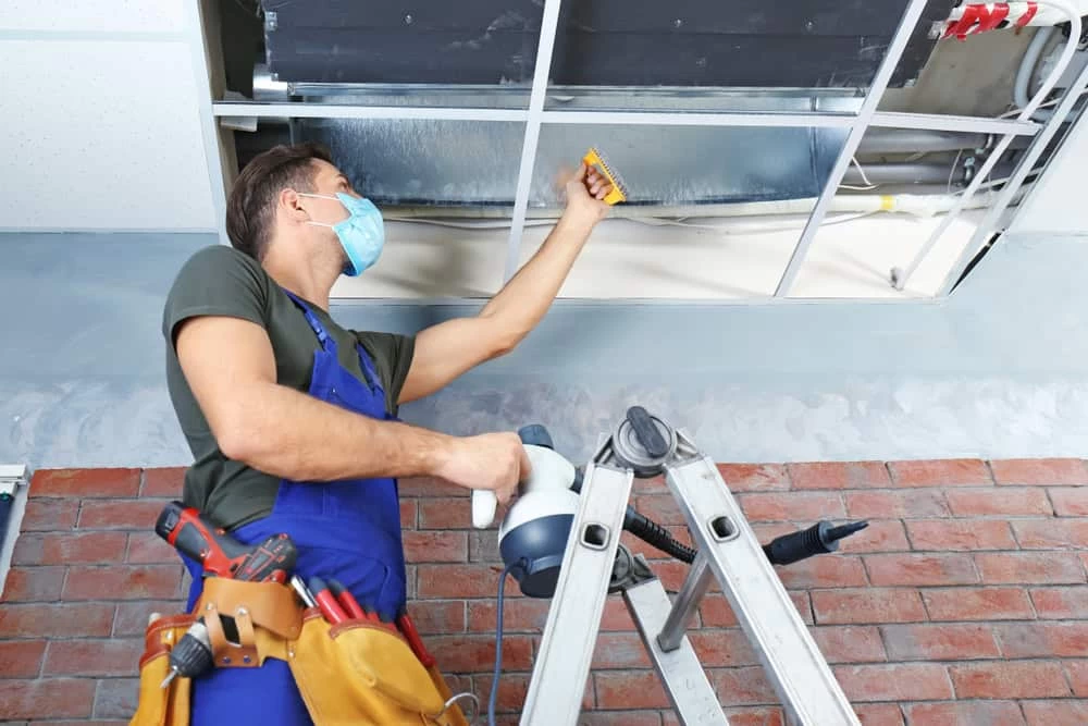 A maintenance worker wearing a mask and tool belt stands on a ladder, inspecting and repairing exposed ductwork in a ceiling&mdash;ideal for air duct services New Jersey or dryer vent cleaning New Jersey.