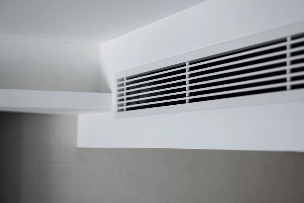 A close-up view of a modern white air conditioning vent set into a white ceiling and wall, with horizontal slats and a grey wall below&mdash;perfect for showcasing professional air duct services New Jersey.