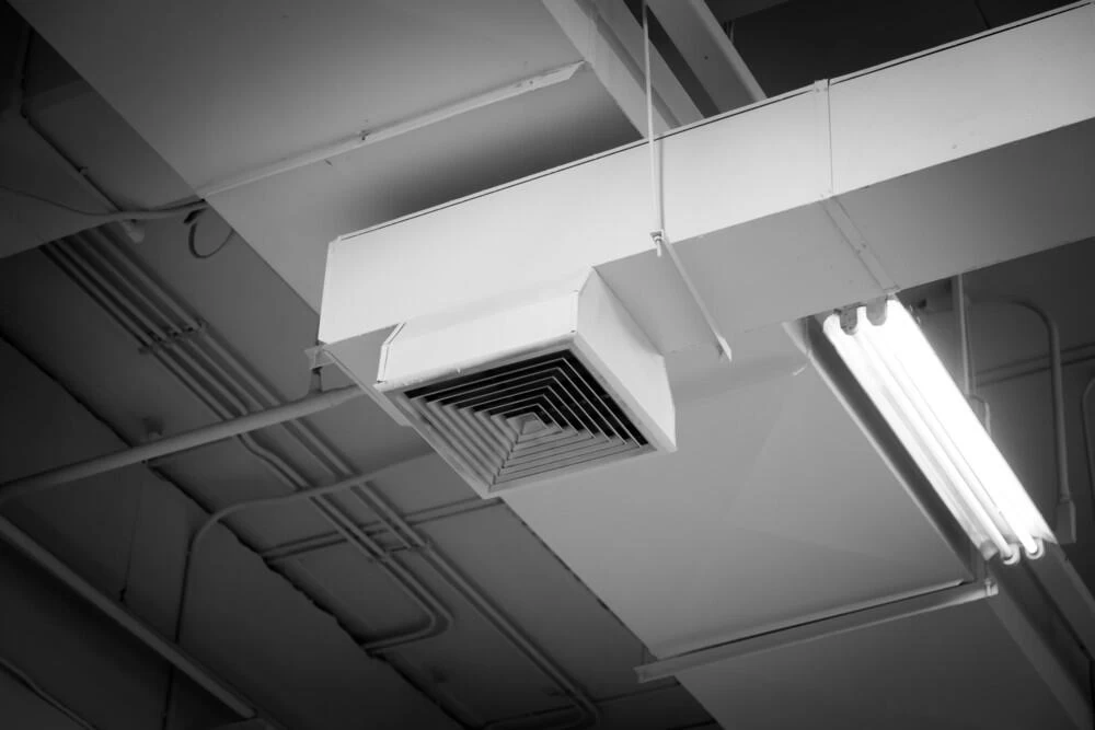 A black and white photo showing a ceiling with an air vent, visible pipes, and a fluorescent light fixture&mdash;highlighting industrial design elements and geometric lines reminiscent of spaces serviced by air duct services New Jersey.