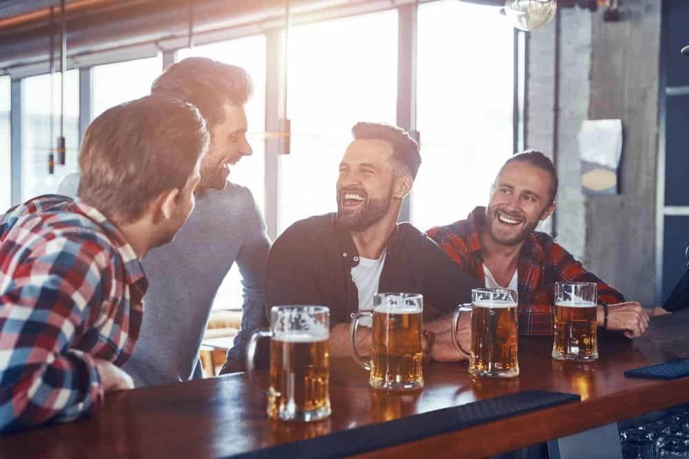 Four men sit at a bar, smiling and laughing together over large glasses of beer. The casual, sunlit setting has fresh air circulating&mdash;like after expert air duct services New Jersey brings to homes and businesses.