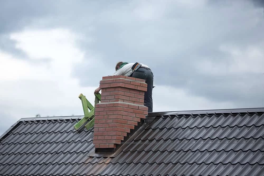 A person is repairing or inspecting a red brick chimney on a dark metal roof under a cloudy sky, ensuring safety much like thorough dryer vent cleaning New Jersey experts provide.