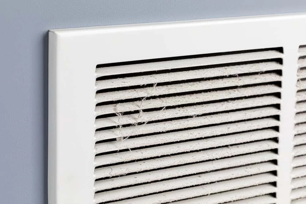 A close-up of a white air vent cover mounted on a gray wall, showing a buildup of dust and cobwebs on the vent slats&mdash;a reminder of the importance of regular air duct services New Jersey homeowners need.